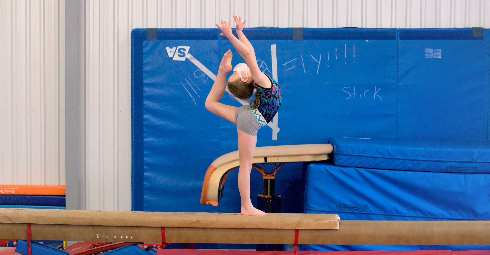 A young gymnast practicing a ring pose on a balance beam at Gymworld Adventures in Gymnastics, a recreational and competitive gymnastics facility in Northwest London