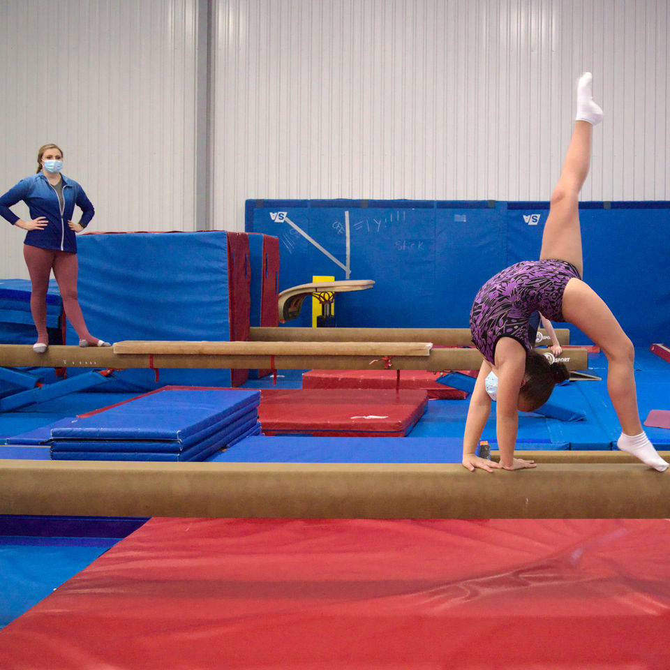 Coach looks on as young gymnast on a balance beam practices a back walkover at Gymworld Adventures in Gymnastics facility in North London.